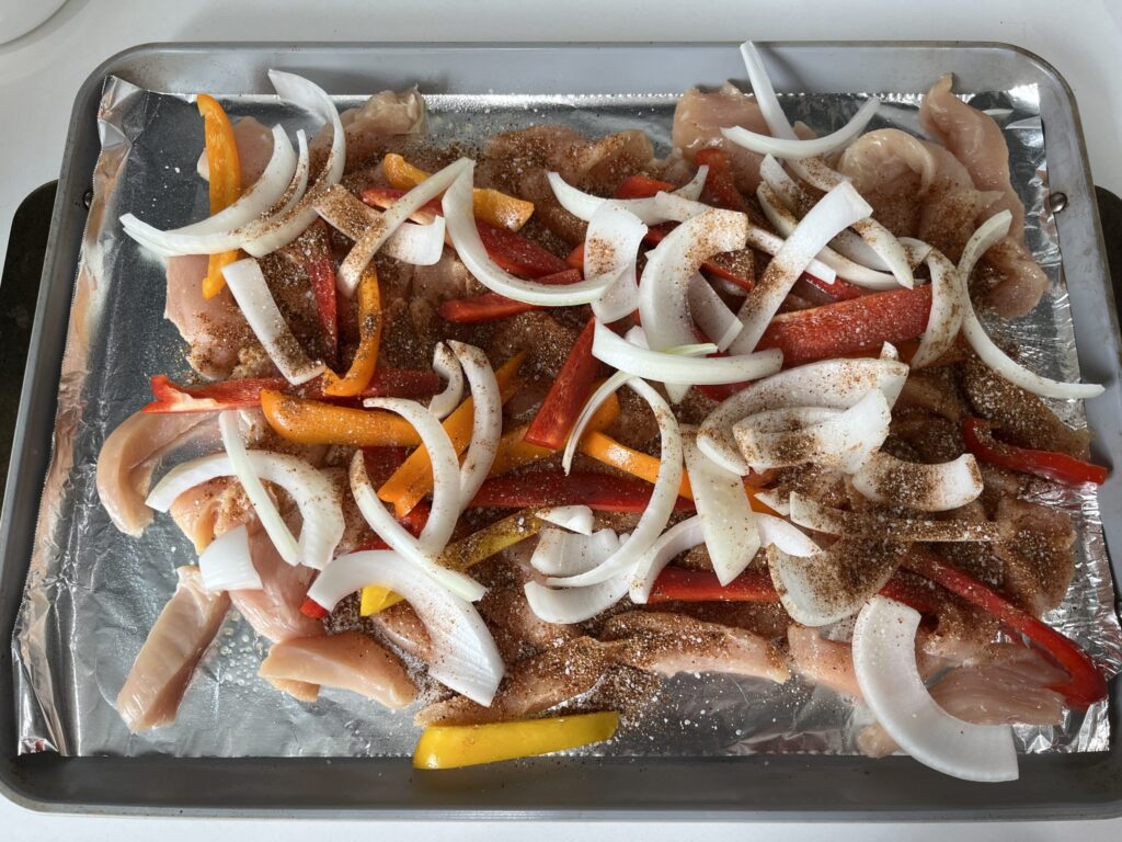 Raw chicken, onions, peppers, and spices mixed together on a baking sheet.