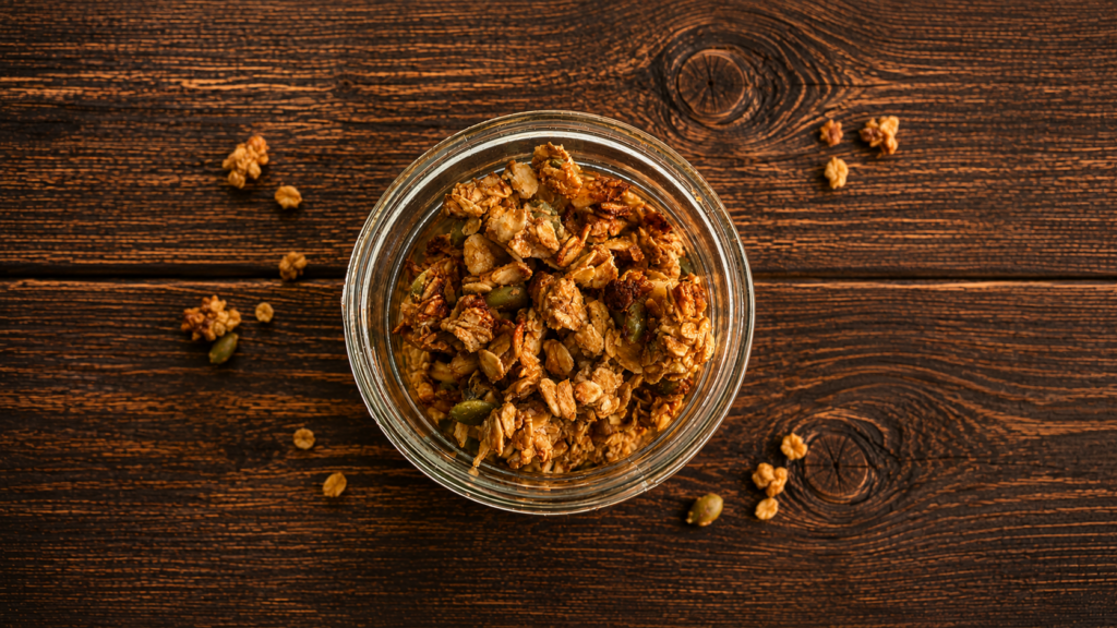 An open jar of high protein granola with granola clusters scattered around it. 