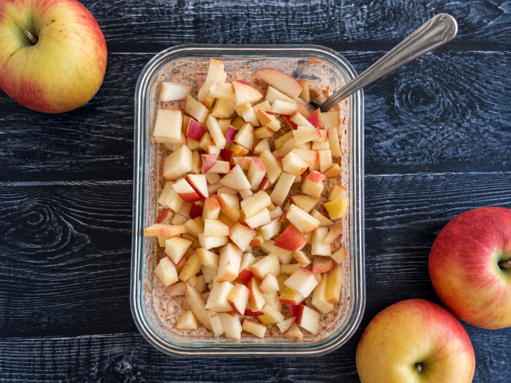 A container of apple overnight oats with a spoon sticking out, three apples scattered around the container.