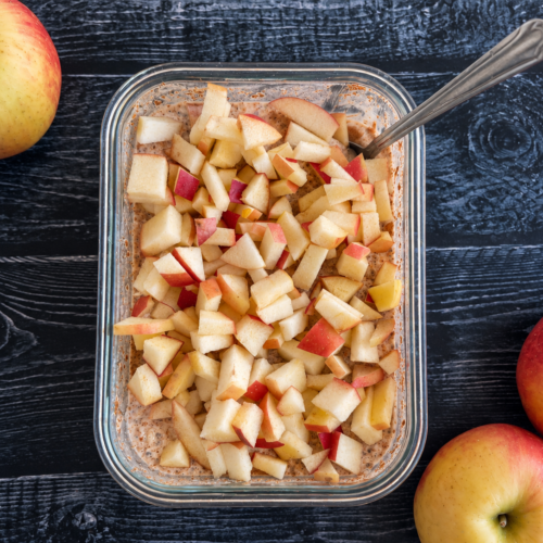 A container of apple overnight oats with a spoon sticking out, three apples scattered around the container.